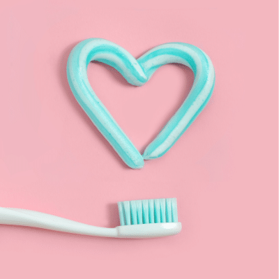 Photo of a blue and white toothbrush on a pink background with aqua blue toothpaste in the shape of a heart above it, representing when to replace your toothbrush
